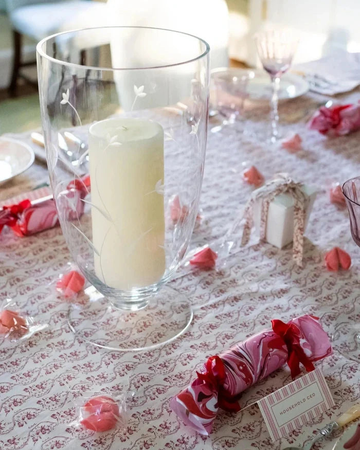 Decorative table setting with a candle in a glass holder, pink paper crackers, and a patterned tablecloth.